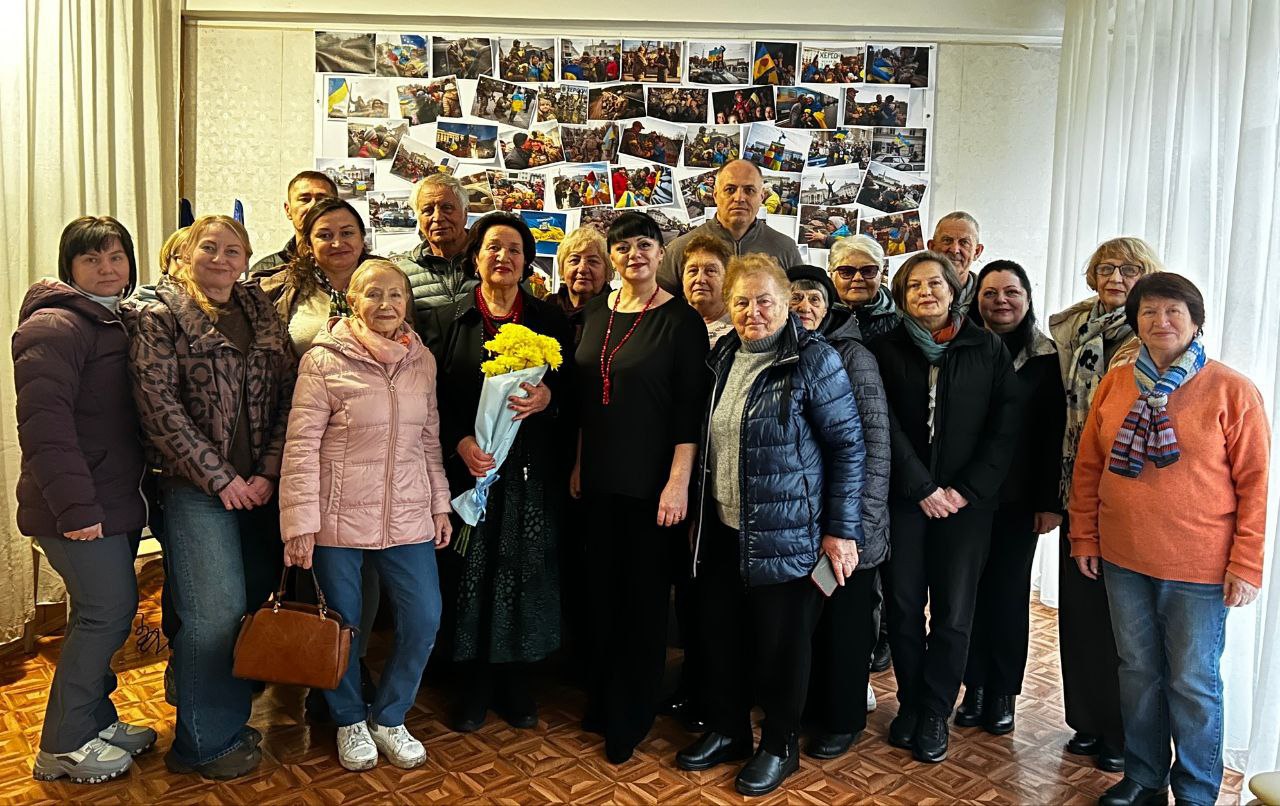 Групова фотографія, на якій стоять приблизно 18 осіб, переважно жінки, у приміщенні. У центрі група людей тримає букет жовтих квітів. На задньому плані видно стіну, повністю вкриту невеликими фотографіями, пов'язаними з Україною.