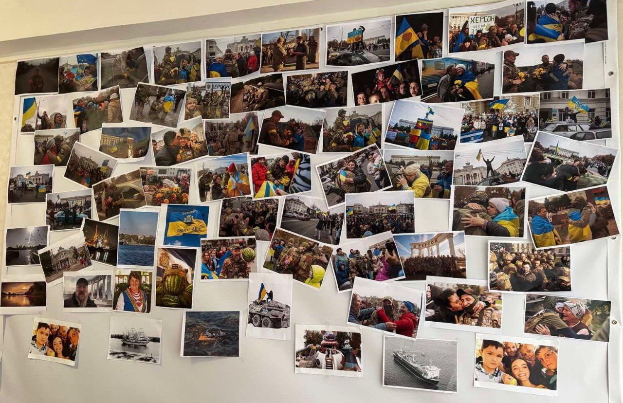 Велика стіна, повністю вкрита безліччю роздрукованих фотографій, накладених одна на одну. Більшість знімків відображають сцени радості та натовпів людей, часто з українськими прапорами та військовими, а також кілька фотографій пейзажів, сімейних моментів та військової техніки.