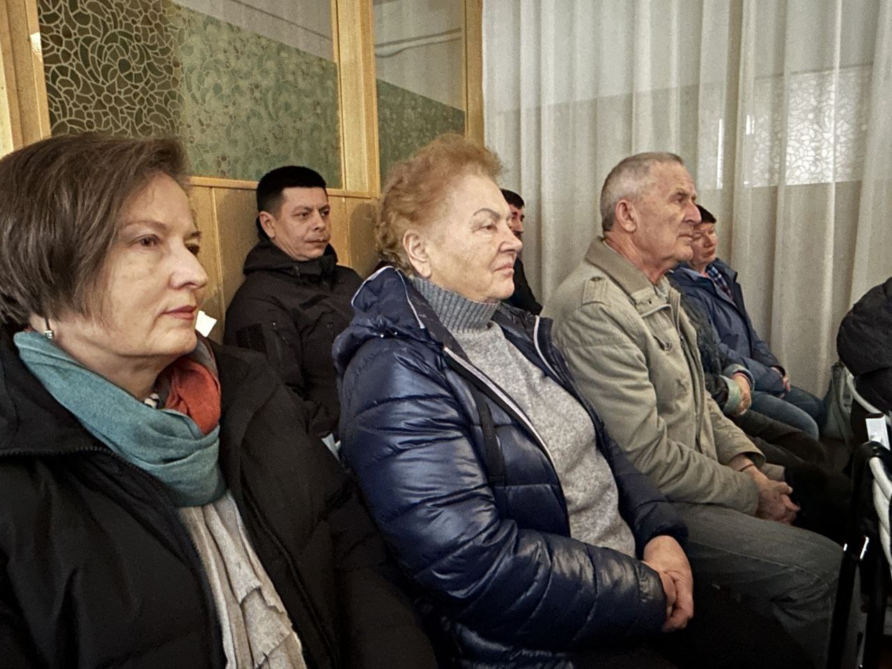 Група людей похилого віку та середнього віку сидять в аудиторії або на зборах. На передньому плані видно трьох осіб у зимовому або демісезонному одязі, які уважно слухають. Знімок зроблено збоку, акцент на глядачах.