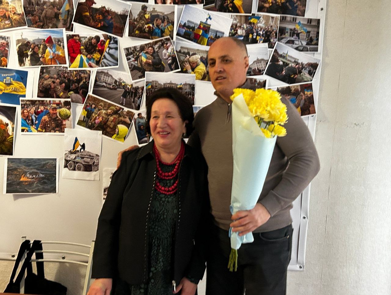 Жінка з червоним намистом і усміхнений чоловік, який тримає великий букет жовтих квітів, позують поруч. Вони стоять на тлі стіни, повністю вкритої невеликими фотографіями, пов'язаними з Україною, включаючи кадри з прапорами та військовою технікою.