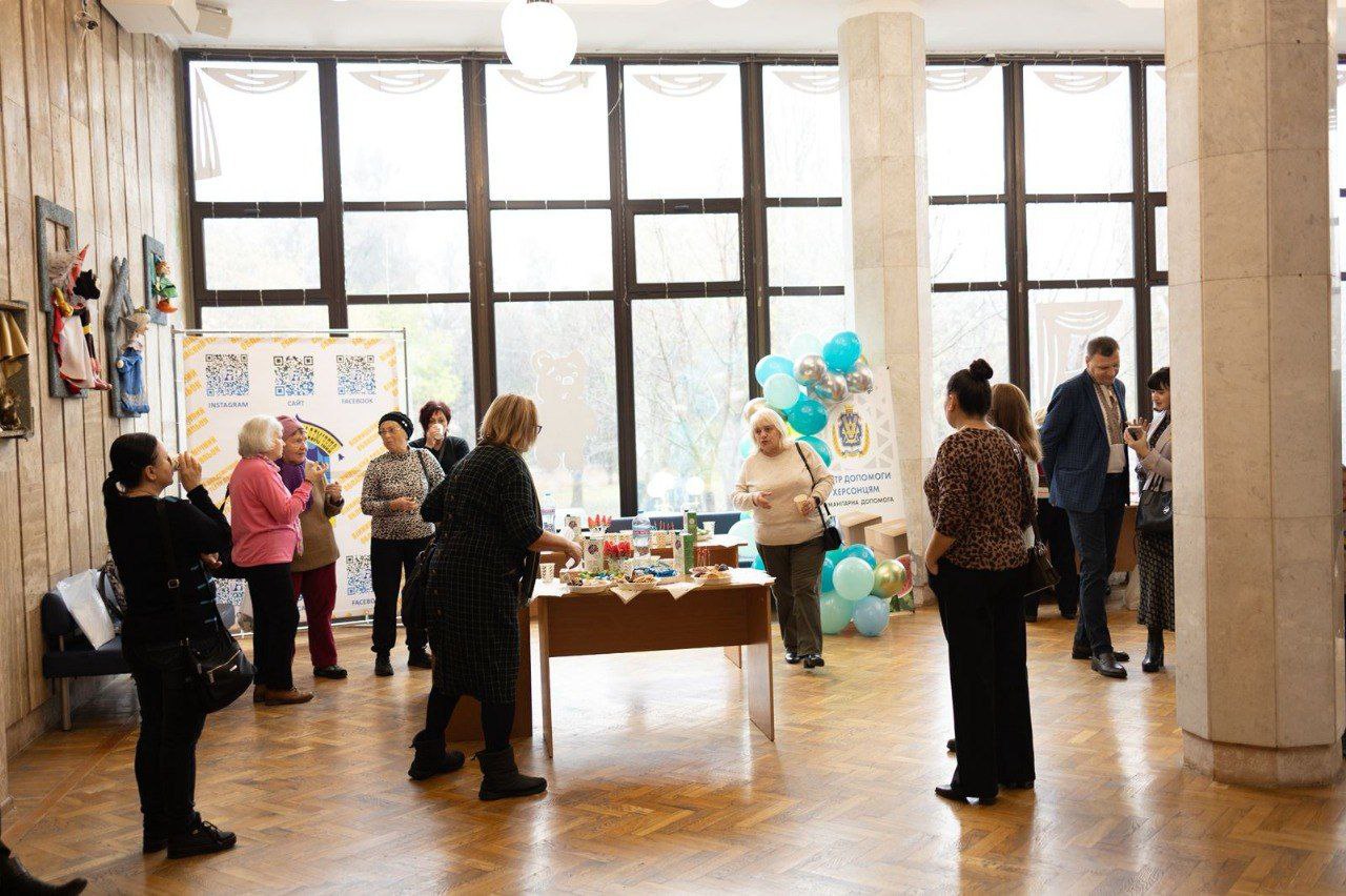 Група людей на зібранні чи заході у світлому просторому приміщенні з великими вікнами та паркетною підлогою. У центрі розташований стіл з предметами для продажу або частування. Ліворуч жінки розглядають стіну з виробами, а праворуч видно повітряні кульки та стенд з інформацією про благодійну чи соціальну подію.