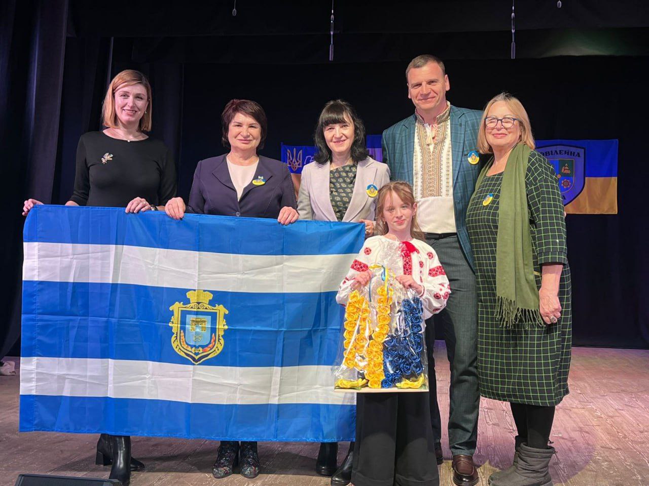 П'ятеро дорослих і одна дівчинка стоять на сцені, тримаючи великий синьо-білий прапор із гербом у центрі (прапор міста Херсона). Чоловік одягнений у вишиванку, а дівчинка — у вишиту блузку. На фоні видно банер із частиною напису та українською символікою. Учасники мають прикріплені синьо-жовті символи, демонструючи підтримку України.