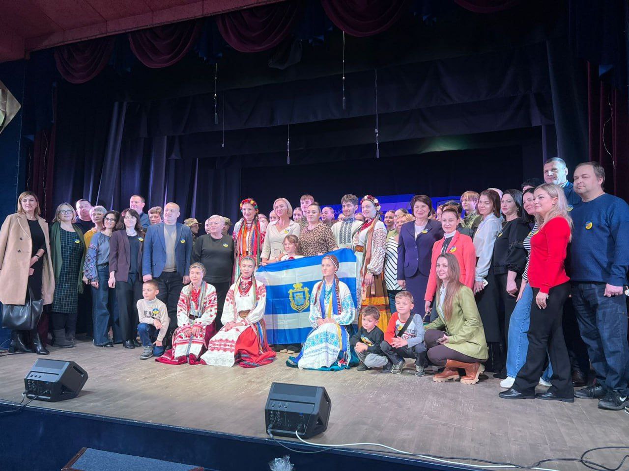 Велика група людей, серед яких дорослі та діти, стоять та сидять на сцені. Деякі учасники одягнені в традиційний український одяг (вишиванки та вінки). В центрі діти тримають прапор, імовірно, прапор міста Херсона, оскільки він синьо-білий із гербом. Загальна атмосфера святкова та патріотична.