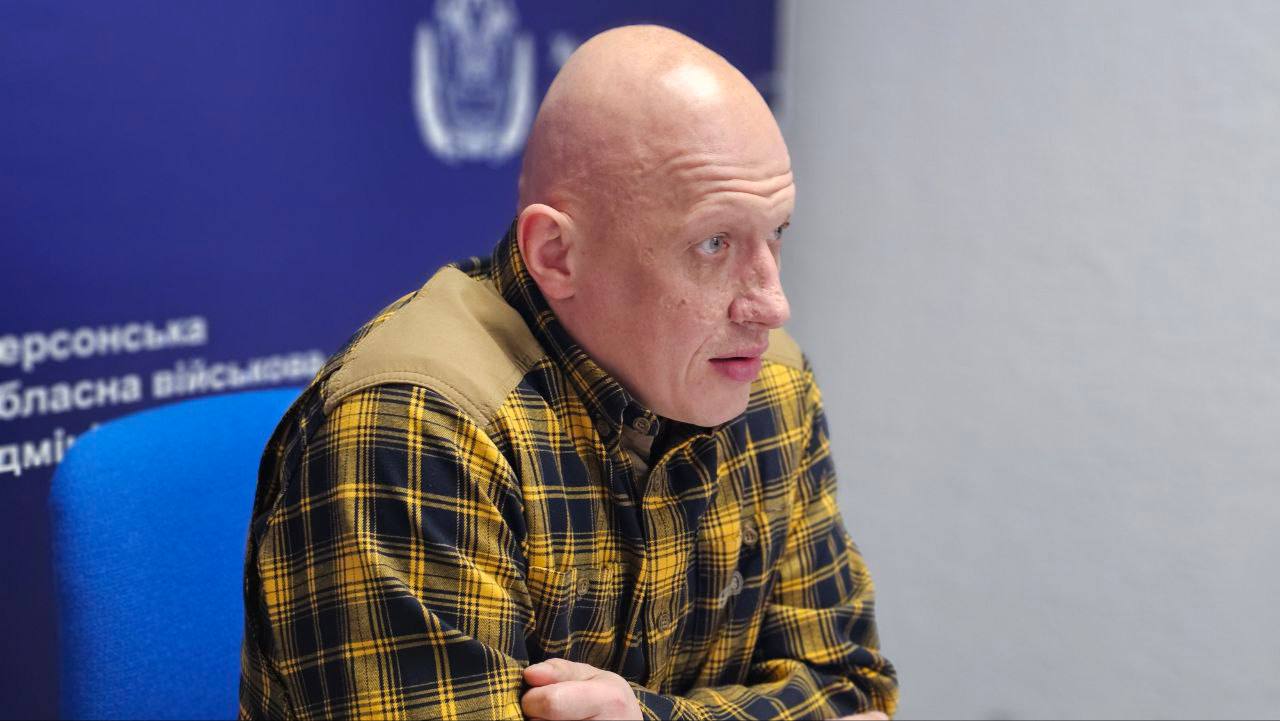 Портретний знімок великим планом лисого чоловіка в жовто-чорній картатій сорочці. Чоловік сидить на синьому стільці й дивиться вправо. На задньому плані розташований синій банер із логотипом Херсонська обласна військова адміністрація.