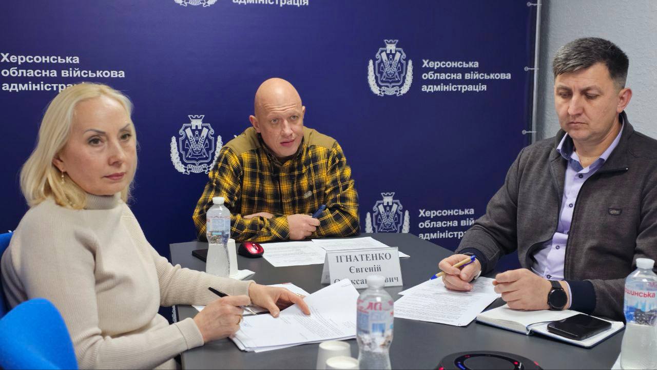 Троє людей – дві жінки і чоловік – сидять за столом на діловій зустрічі. Жінка на передньому плані та чоловік праворуч дивляться на документи, а чоловік у центрі, одягнений у жовто-чорну сорочку, дивиться вперед. На задньому плані розташований синій банер із логотипом Херсонська обласна військова адміністрація. На столі видно пляшки з водою.