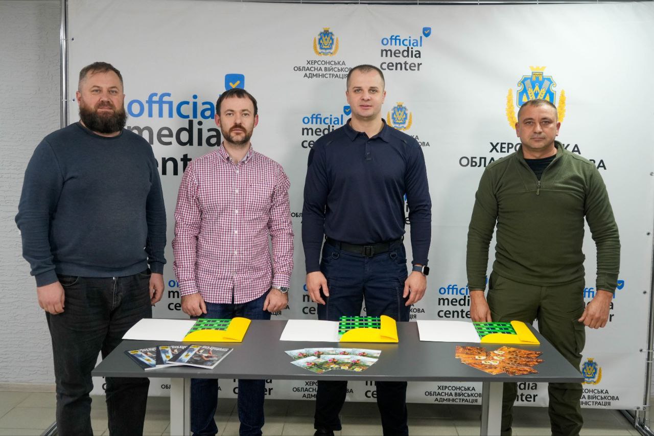 Четверо чоловіків стоять за столом перед пресволом, на якому багаторазово зображено логотип Херсонська обласна військова адміністрація та official media center. На столі лежать друковані матеріали та жовто-зелені папки. Чоловіки одягнені у повсякденний та військовий одяг.