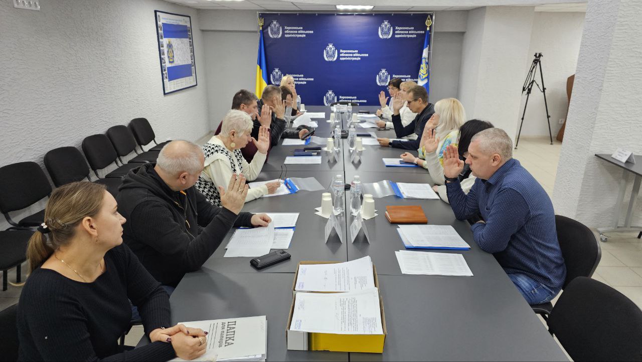 Група людей (близько 14 осіб) сидить за довгим столом під час засідання. Кілька учасників, розташованих по обидва боки столу, підняли праві руки для голосування чи присяги. На стіні позаду видно банери з емблемою Херсонської обласної військової адміністрації та українським прапором.