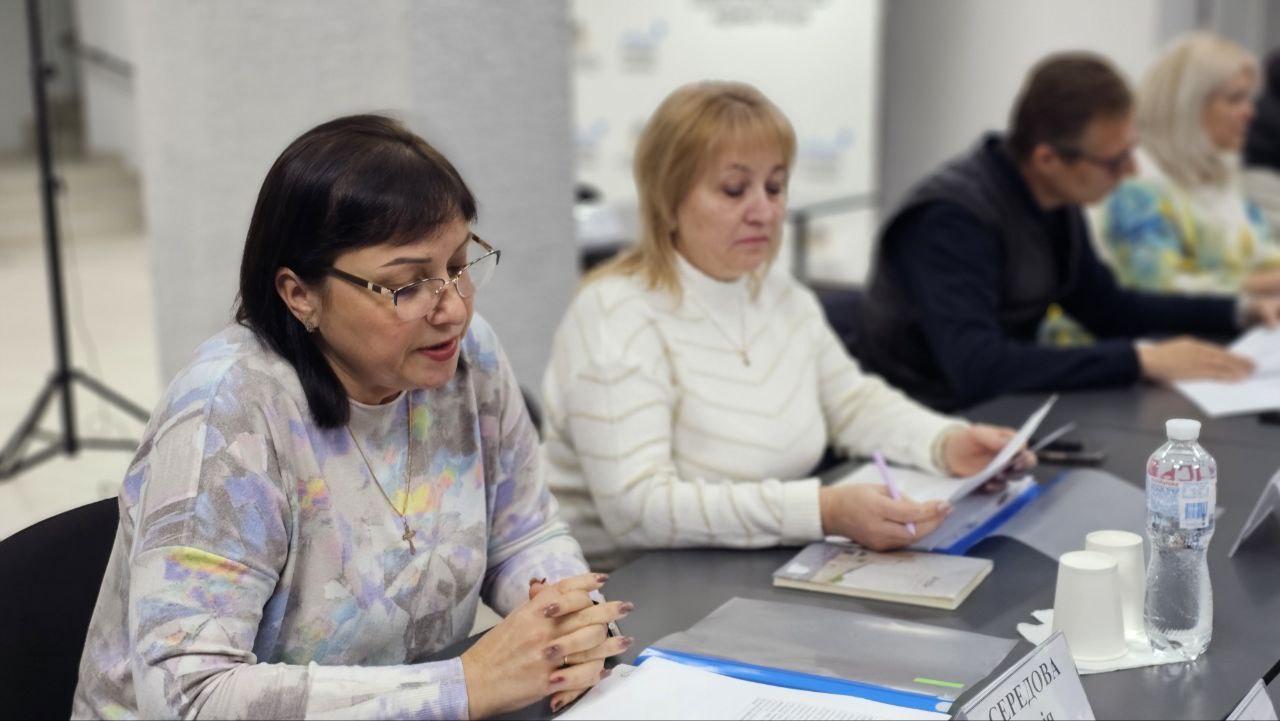 Крупний план двох жінок, які беруть участь у засіданні за столом. Жінка на передньому плані (зліва) у світлому светрі та окулярах, склала руки перед собою, можливо, говорить. Інша жінка (по центру) у білому светрі читає документи. На столі видно папери та пляшка води.