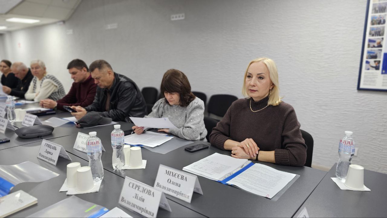 Крупний план учасників засідання за столом. На передньому плані справа сидить жінка зі світлим волоссям у темно-коричневому светрі, дивлячись прямо. Перед нею лежать документи та іменна табличка. Зліва від неї сидять інші учасники, включно з жінкою, що читає документи, та чоловіками, а також видно іменні таблички.