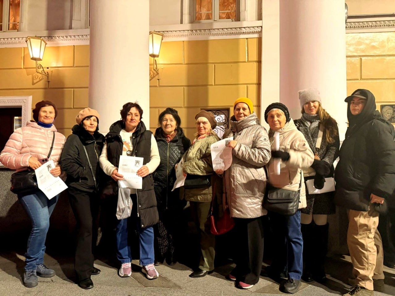 Групова фотографія дев'яти жінок у зимовому одязі, які стоять вночі перед фасадом будівлі з колонами та ліхтарями. Кілька жінок тримають у руках папери або сертифікати.