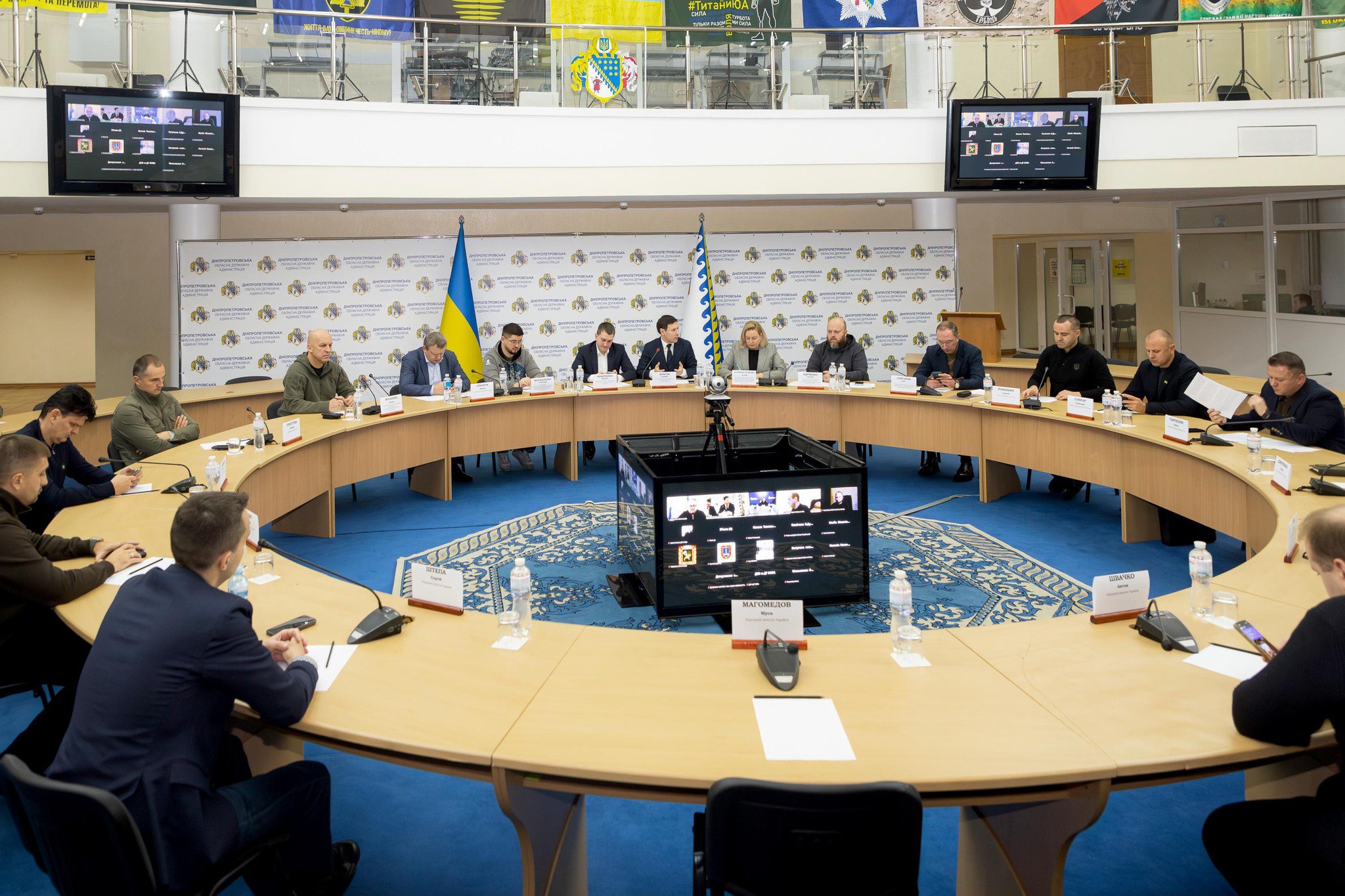 Нарада у великому конференц-залі, де близько 20 осіб, переважно чоловіків, сидять за круглим столом. У центрі столу розташований монітор для відеозв'язку. На стіні позаду видно український прапор та банери з емблемами Херсонської обласної військової адміністрації. На двох екранах по обидва боки зали відображаються учасники онлайн-конференції.