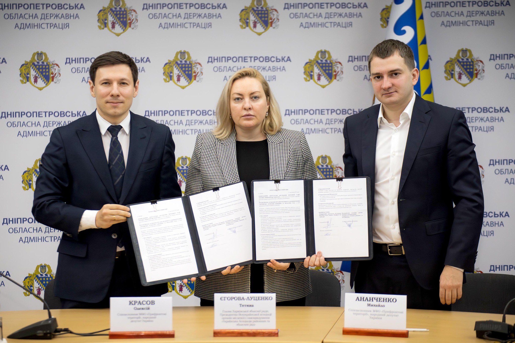 Троє людей (двоє чоловіків у костюмах і жінка по центру в чорно-білому піджаку) стоять за столом і демонструють підписані документи. На задньому плані видно банери з написом: ДНІПРОПЕТРОВСЬКА ОБЛАСНА ДЕРЖАВНА АДМІНІСТРАЦІЯ. Перед ними стоять іменні таблички.