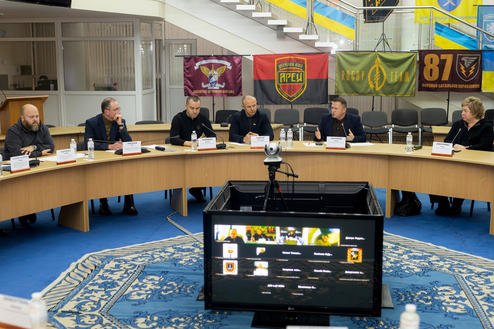 Нарада семи осіб (шість чоловіків та одна жінка) за напівкруглим столом у конференц-залі. На столі видно іменні таблички. Перед столом розташовано великий монітор для відеоконференції. На задньому плані, під сходами, висять численні військові прапори та прапори України.