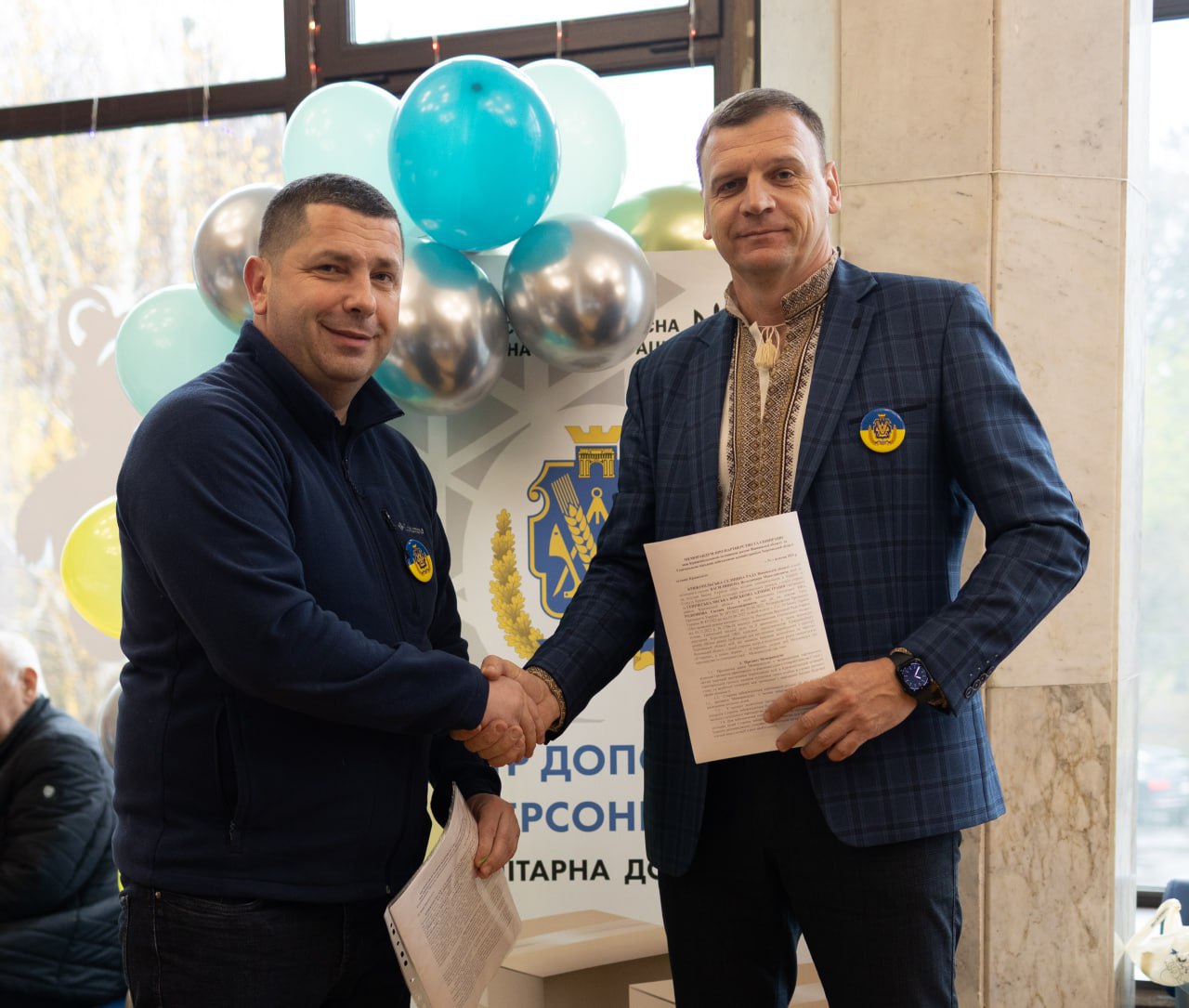 Двоє чоловіків потискають один одному руки, стоячи перед стендом із логотипом Херсонської обласної адміністрації та повітряними кульками блакитного, жовтого та срібного кольорів. Чоловік праворуч одягнений у вишиванку під синім піджаком і тримає в руці документ.