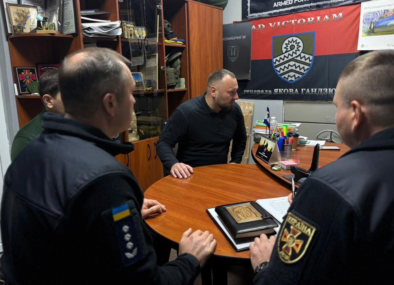 Четверо чоловіків у темній формі/одязі сидять навколо круглого столу в офісному приміщенні. Один із чоловіків, що сидить ближче до центру, тримає розгорнуту чорну книжку чи теку. На стіні позаду висять військові банери з емблемами та гаслами.