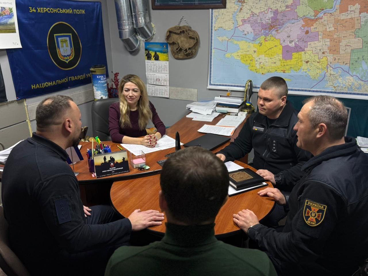 Чотири особи (одна жінка зі світлим волоссям у бордовому светрі та троє чоловіків у темній формі/одязі) сидять за круглим столом в офісі. Чоловіки дивляться на жінку, а один із них тримає закриту чорну книжку чи теку.