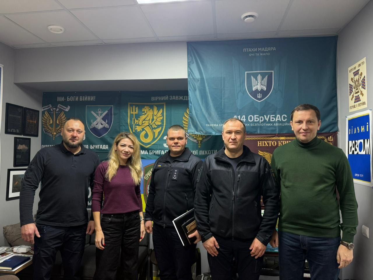 Групова фотографія п'яти людей (чотирьох чоловіків у темному одязі та жінки зі світлим волоссям у бордовому светрі), які стоять в офісному приміщенні на тлі великих банерів військової тематики.