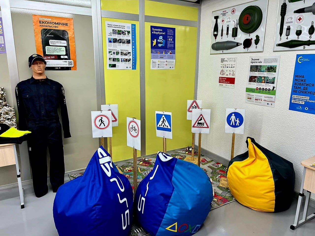 Навчальний клас із цивільного захисту та безпеки дорожнього руху. У кімнаті розміщені кольорові жовто-зелені панелі. Зліва стоїть манекен у поліцейській формі. На підлозі лежить килимок із дорожнім малюнком, на якому стоять три кольорові м'які крісла-мішки. Навколо них розташовані дорожні знаки. На стінах висять навчальні плакати, включно з інформацією про мінну небезпеку.