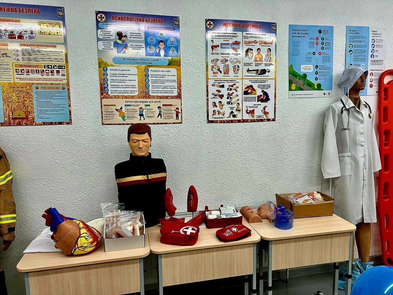 Навчальний клас або куток із плакатами на стіні, присвяченими різним аспектам безпеки: Психологічна безпека, Медична допомога, а також правилам, як поводитися в небезпечних ситуаціях. На столах розташовані навчальні манекени, медичні приналежності, зокрема модель серця, аптечки та манекен для надання першої допомоги. Справа стоїть манекен у білому халаті медсестри/лікаря.