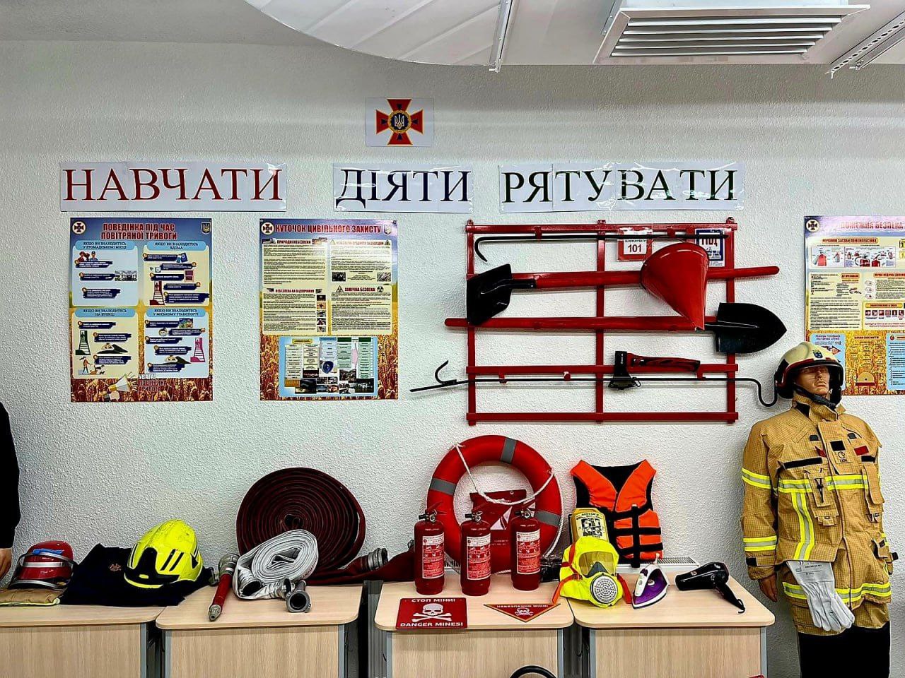 Навчальний клас або куток, присвячений цивільному захисту, пожежній та мінній безпеці. На стіні розміщені плакати з написами: НАВЧАТИ, ДІЯТИ, РЯТУВАТИ та схеми правил поведінки. На червоній стійці висить пожежний інвентар (сокира, лом, лопата). На столі представлені засоби пожежогасіння та рятування: пожежні шоломи, рукав, вогнегасники, рятувальний круг, жилет та протигази. Зліва стоїть манекен у пожежному костюмі.