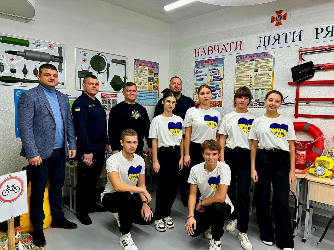 Групова фотографія дорослих (чоловіків) та підлітків (хлопців та дівчат), що стоять у навчальному класі. Підлітки одягнені в білі футболки з синьо-жовтим серцем та написом: ЖИВИ. На стінах розвішані плакати та інструкції з безпеки та цивільного захисту з написами: НАВЧАТИ, ДІЯТИ, РЯТУВАТИ.