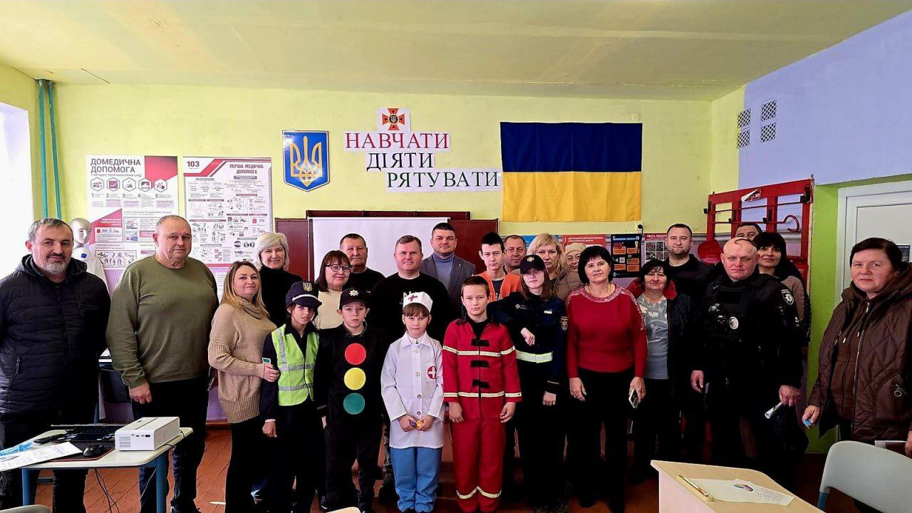 Групова фотографія дорослих і дітей у приміщенні з українським прапором та плакатами про безпеку на стіні. У центрі стоять діти у формі, що імітує пожежників, медиків і працівників дорожніх служб, а навколо них — дорослі вчителі, інструктори та, ймовірно, батьки. Напис на стіні: НАВЧАТИ ДІЯТИ РЯТУВАТИ.