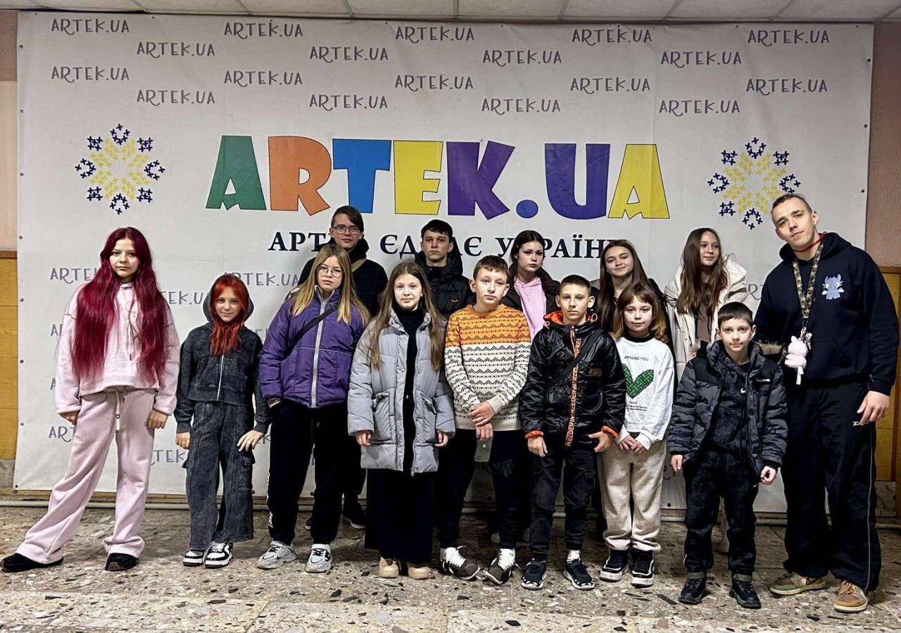 Група підлітків (близько 13 дітей) і один дорослий чоловік стоять разом, позуючи перед великим банером. На банері написано: ARTEK.UA великими кольоровими літерами та гасло АРТЕК – ЦЕ УКРАЇНА. Стіна за ними також прикрашена повторюваними написами: ARTEK.UA.