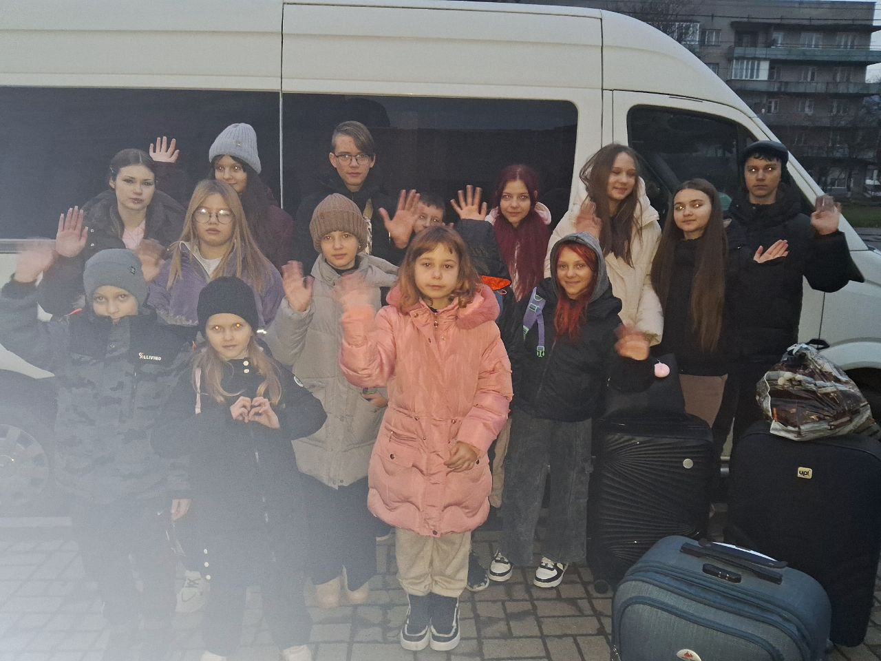 Група підлітків стоїть біля білого мікроавтобуса, більшість з них махають руками на камеру. Діти вдягнені у зимовий або демісезонний одяг. Поруч стоять кілька великих валіз, що свідчить про подорож.