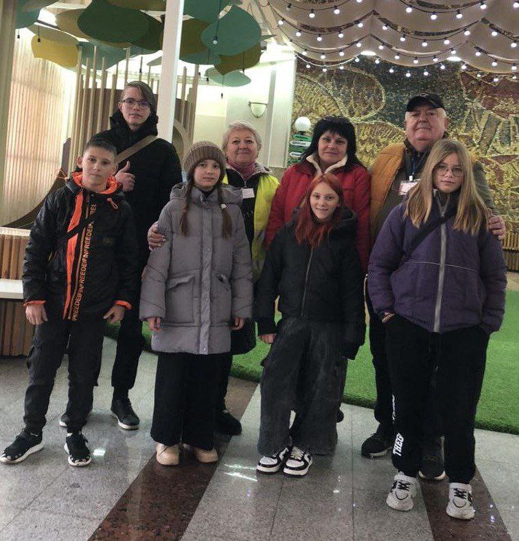 Група з семи людей, включаючи чотирьох підлітків та трьох дорослих, стоять разом у приміщенні з мозаїчною стіною на задньому плані. Підлітки та двоє дорослих одягнені в теплий одяг. Приміщення має мармурову підлогу та декоративну стелю.