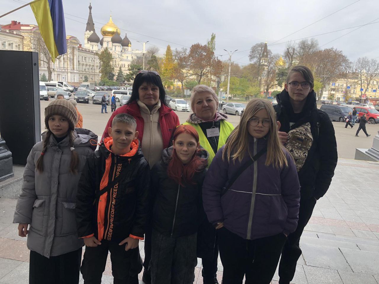 Група з шести підлітків та двох дорослих жінок стоять на сходах на відкритому повітрі. На задньому плані видно площу, автомобілі та будівлю з золотими куполами, ймовірно, собор або церкву. Ліворуч також видно український прапор.