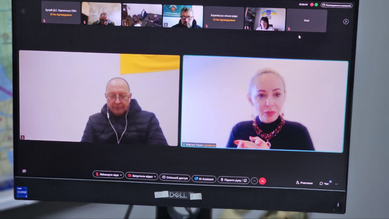 Монітор комп'ютера під час відеоконференції. На екрані видно кількох учасників у маленьких вікнах угорі, а внизу — двох основних доповідачів: чоловіка в темній куртці на тлі жовто-синього прапора та світловолосу жінку в чорному светрі. Написи містять імена учасників та елементи керування конференцією.