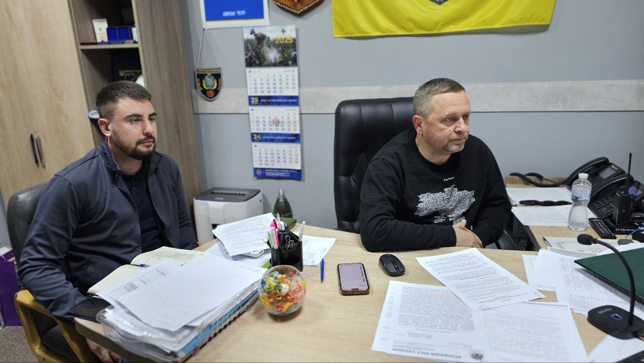 Два чоловіки сидять за робочим столом у кабінеті. Стіл завалений паперами та канцелярським приладдям. Ліворуч сидить молодший чоловік у темній куртці, праворуч – старший чоловік у чорному светрі. На задньому плані видно календар та фрагмент прапора.