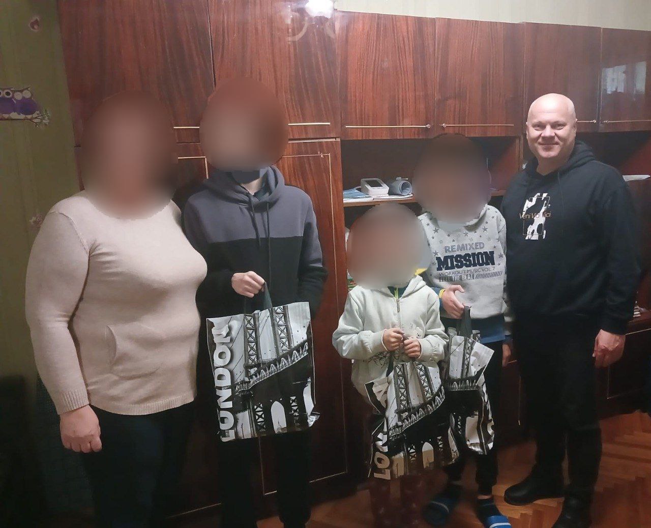 Чоловік, жінка та троє дітей стоять разом у кімнаті перед шафою, тримаючи великі пакети з написом: London.