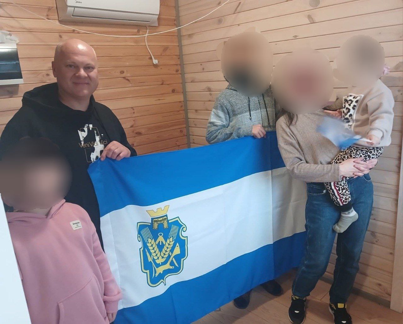 Чоловік та сім'я з трьома дітьми тримають великий синьо-білий прапор Херсонської області на тлі дерев'яної стіни.