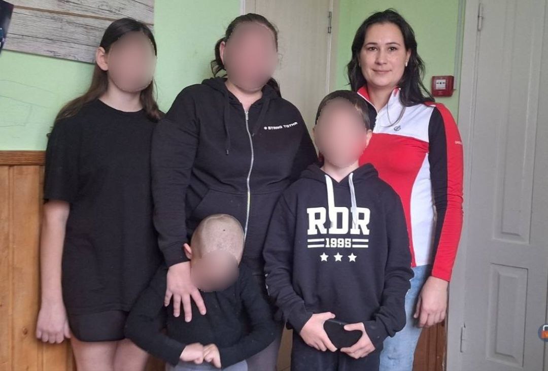 Груповий портрет чотирьох дітей та двох дорослих жінок, які стоять разом у приміщенні на тлі дерев'яної панелі та світлих дверей.