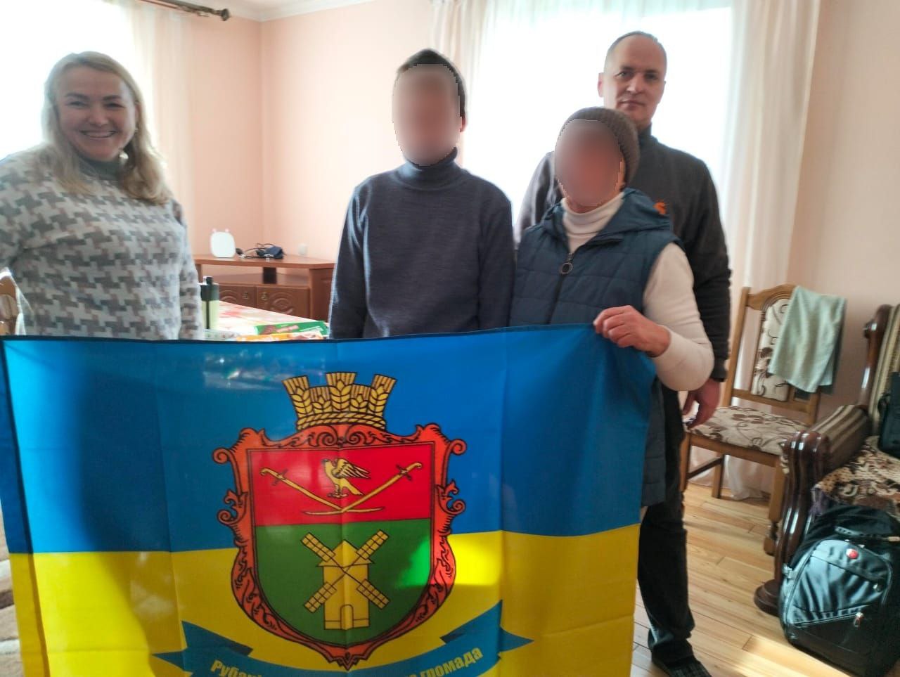 Двоє чоловіків та двоє жінок стоять у приміщенні, тримаючи великий жовто-синій прапор із гербом Рубіжанської міської громади.