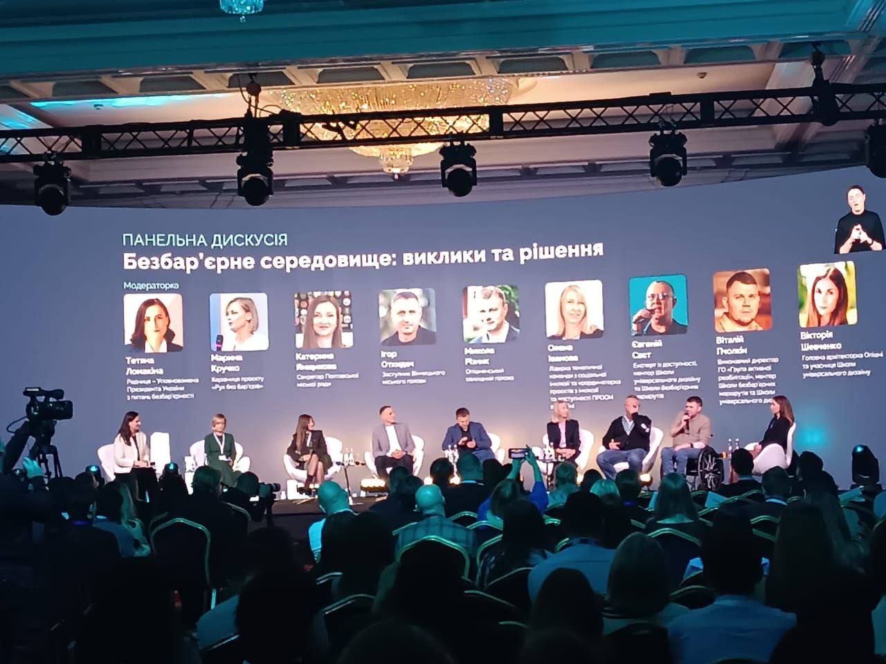 Велика панельна дискусія на конференції, де на сцені сидять дев'ять спікерів перед аудиторією, а на великому екрані відображено тему: Безбар'єрне середовище: виклики та рішення та фотографії її учасників.