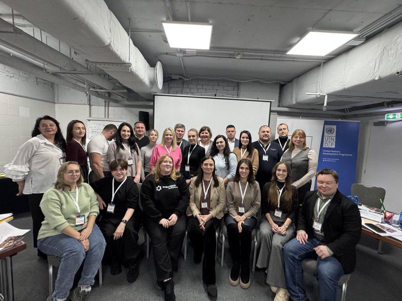 Групова фотографія близько 25 учасників тренінгу або конференції, які стоять і сидять у два ряди перед проекційним екраном, поруч видно банер Програми розвитку ООН (UNDP).