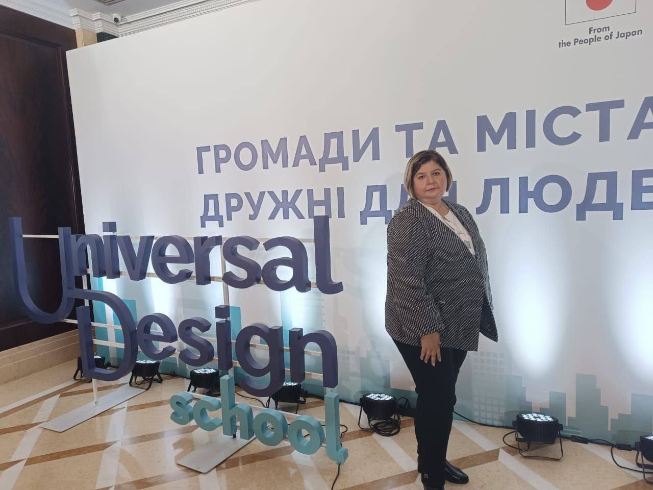 Жінка в чорно-білому піджаку стоїть поруч з великим написом: Universal Design School та банером Громади та міста, дружні для людей на конференції.