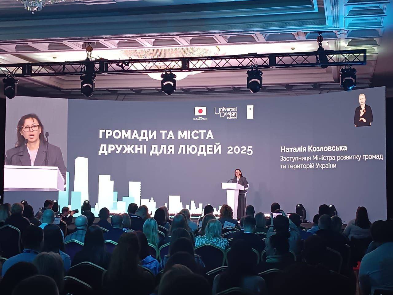 Заступниця міністра розвитку громад та територій України, виступає перед великою аудиторією на конференції під банером Громади та міста, дружні для людей 2025 та логотипом Universal Design School.