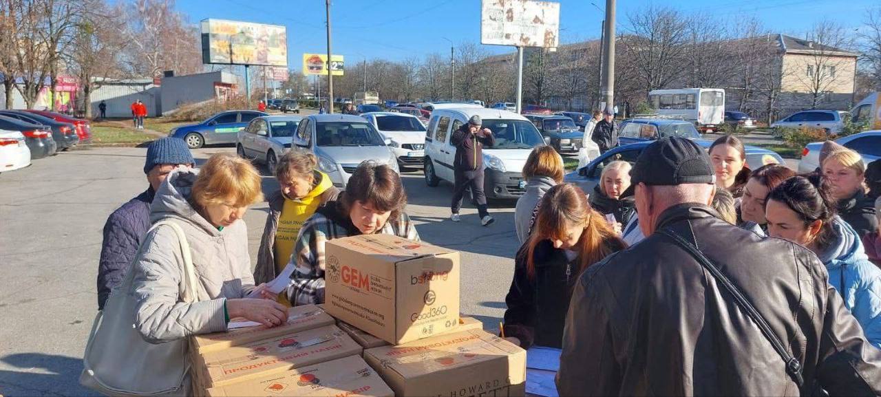 Люди отримують гуманітарну допомогу (коробки з продуктовими наборами, зокрема з логотипом GEM) на відкритій території, схожій на парковку.