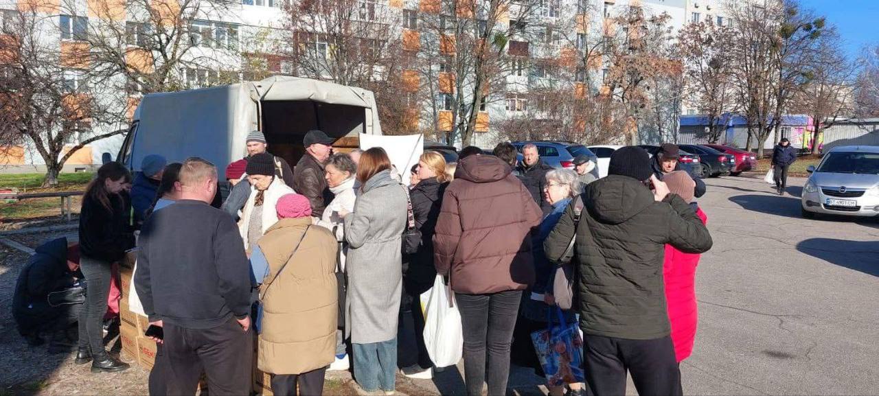 Група людей, які зібралися біля відкритої вантажівки (фургона) для отримання гуманітарної допомоги, на тлі багатоповерхових житлових будинків та парковки.