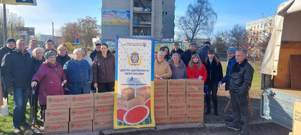 Група людей на вулиці, які стоять біля коробок з гуманітарною допомогою (логотип GEM) та плаката ЦЕНТР ДОПОМОГИ ХЕРСОНЦЯМ від Херсонської ОВА.