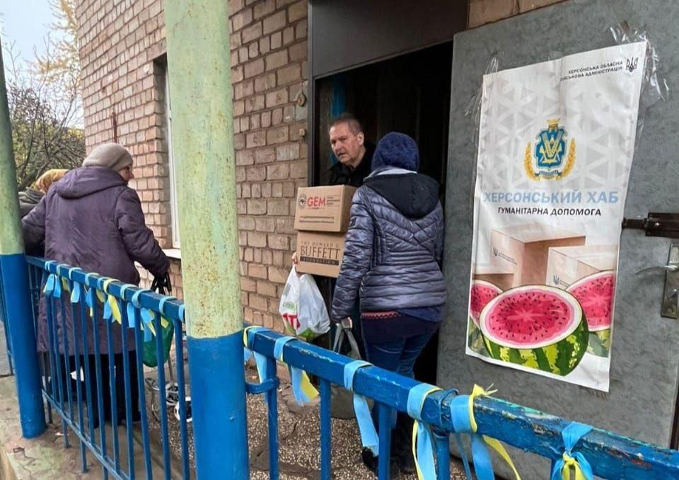Чоловік видає гуманітарну допомогу (коробки GEM/Buffett) двом жінкам біля входу до Херсонського хабу, позначеного банером, на тлі перил, прикрашених синьо-жовтими стрічками.