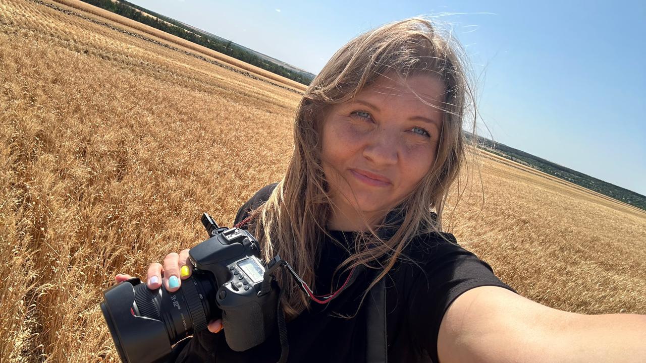 Жінка з фотоапаратом стоїть у пшеничному полі під яскравим сонячним небом.