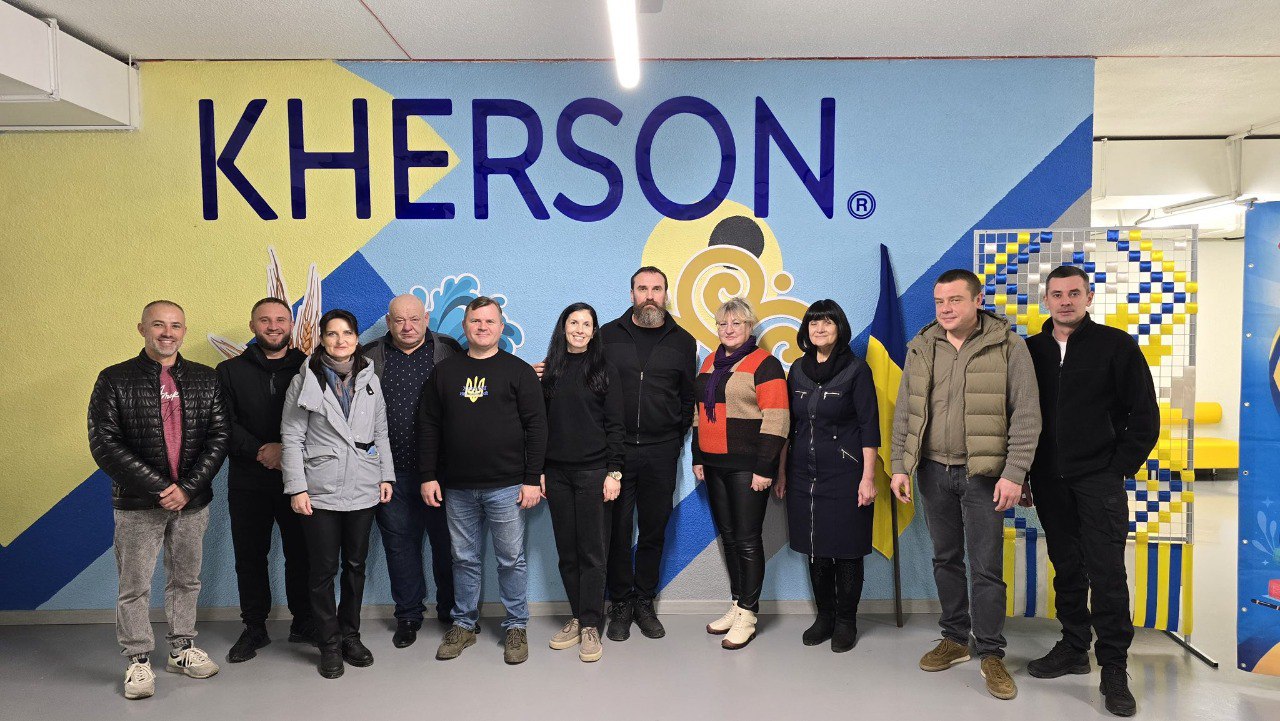 Група з одинадцяти людей стоїть у приміщенні на фоні стіни з великим написом: KHERSON та графічним зображенням у синьо-жовтих тонах. Поруч стоїть український прапор.