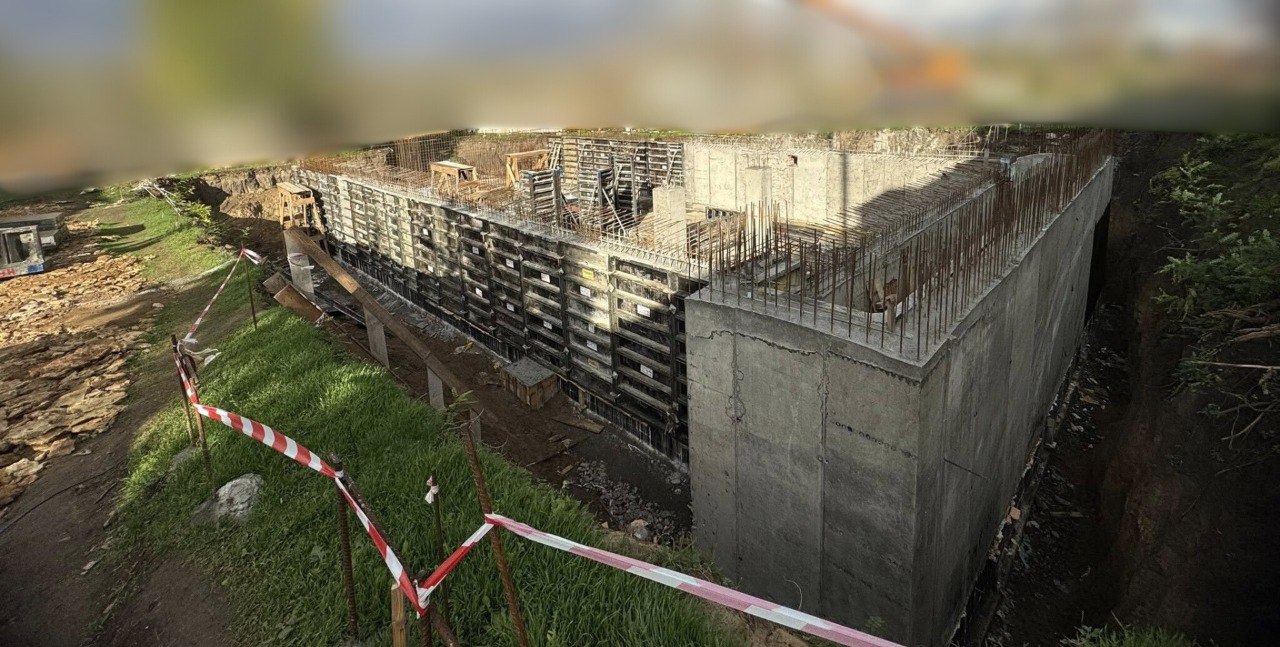 Панорамний знімок демонструє будівництво залізобетонної споруди (можливо, захисного укріплення або фундаменту) з встановленою опалубкою та арматурою, оточеної ґрунтом і травою. Територія обгороджена червоно-білою стрічкою.