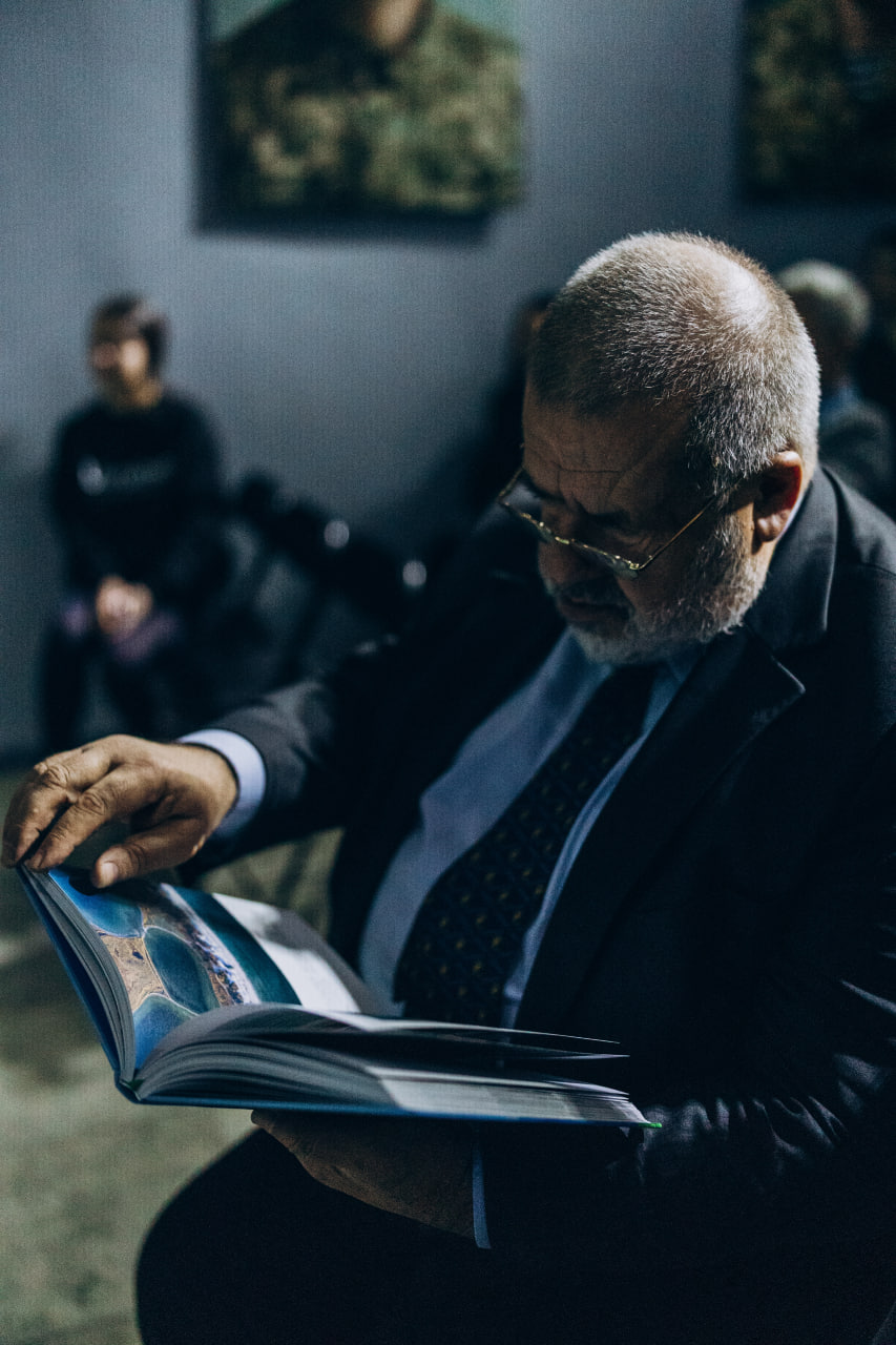 Чоловік у костюмі та окулярах сидить у залі й уважно гортає ілюстровану книгу. На задньому плані розмито видно інших присутніх та картини на стінах.