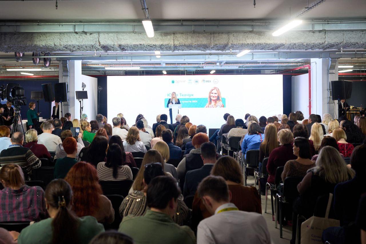 Вид на заповнену аудиторію зі спини: багато людей сидять і слухають спікера (жінку) на сцені. На великому екрані позаду неї видно її зображення та логотипи організацій (зокрема IREX).