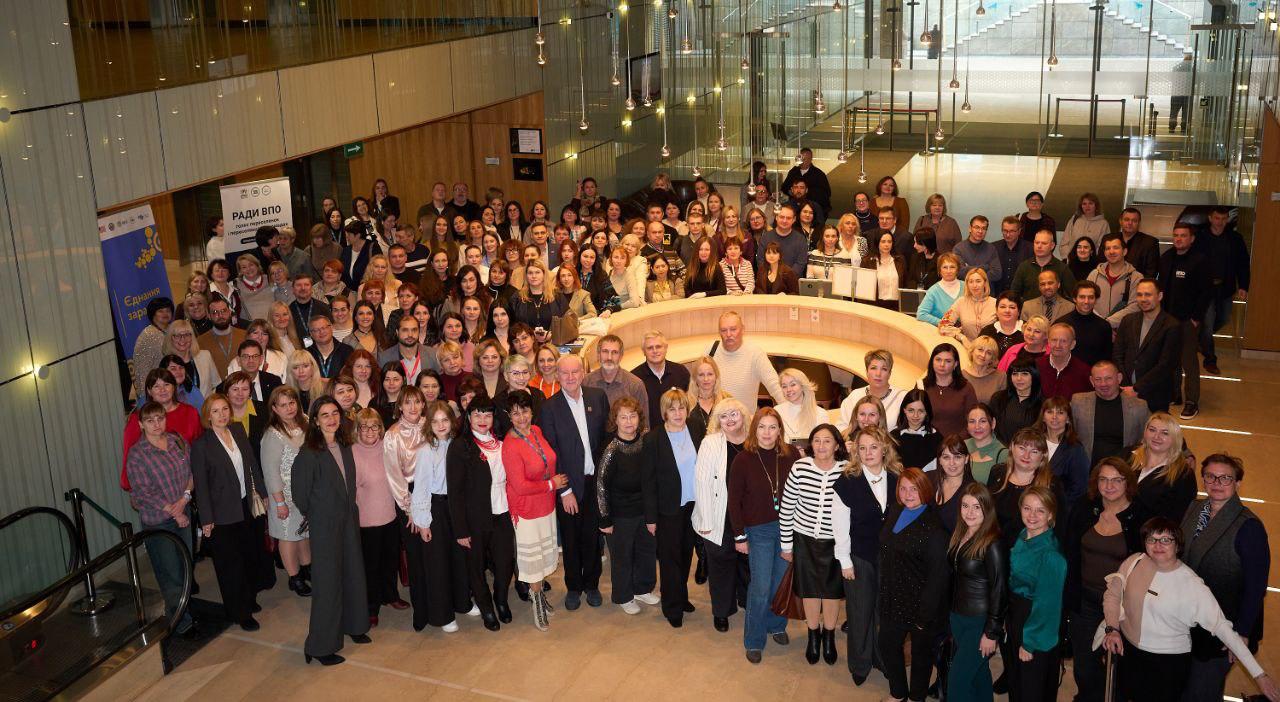 Велика група людей, що складається з чоловіків та жінок різного віку, стоїть і дивиться в камеру, позуючи для спільного фото у світлому сучасному приміщенні. У центрі композиції видно округлу стійку.